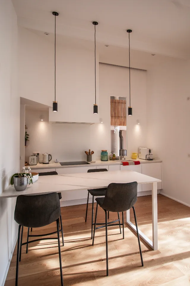 Cuisine blanche avec table haute, trois suspensions noires et spots muraux allumés