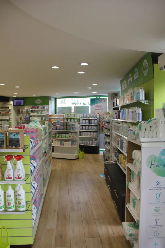 Allée de pharmacie entre rayonnages verts et blancs, spots encastrés allumés au plafond