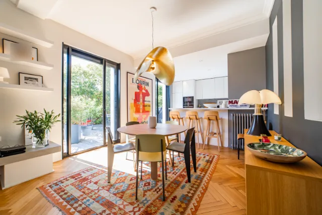 Coin repas avec table ronde, tapis kilim, suspension dorée et cuisine ouverte au fond