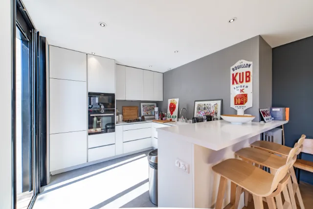 Cuisine blanche sans poignées, fours encastrés, péninsule à tabourets en bois et spots encastrés