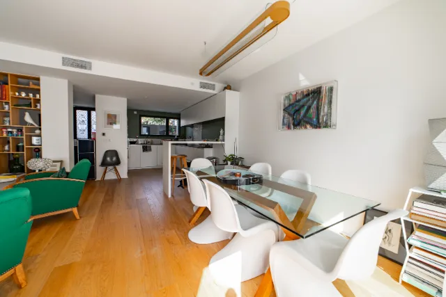 Pièce de vie avec table en verre, chaises blanches, suspension linéaire et cuisine ouverte au fond