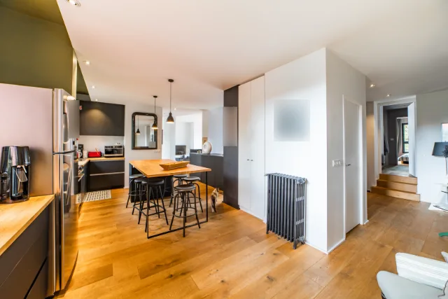 Cuisine ouverte aux façades noires, table haute en bois, deux suspensions et radiateur à colonnes