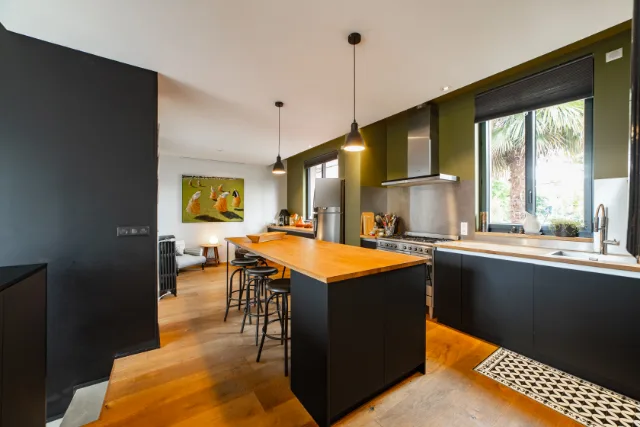Cuisine noire aux murs vert olive, îlot en bois, piano de cuisson, hotte inox et suspensions