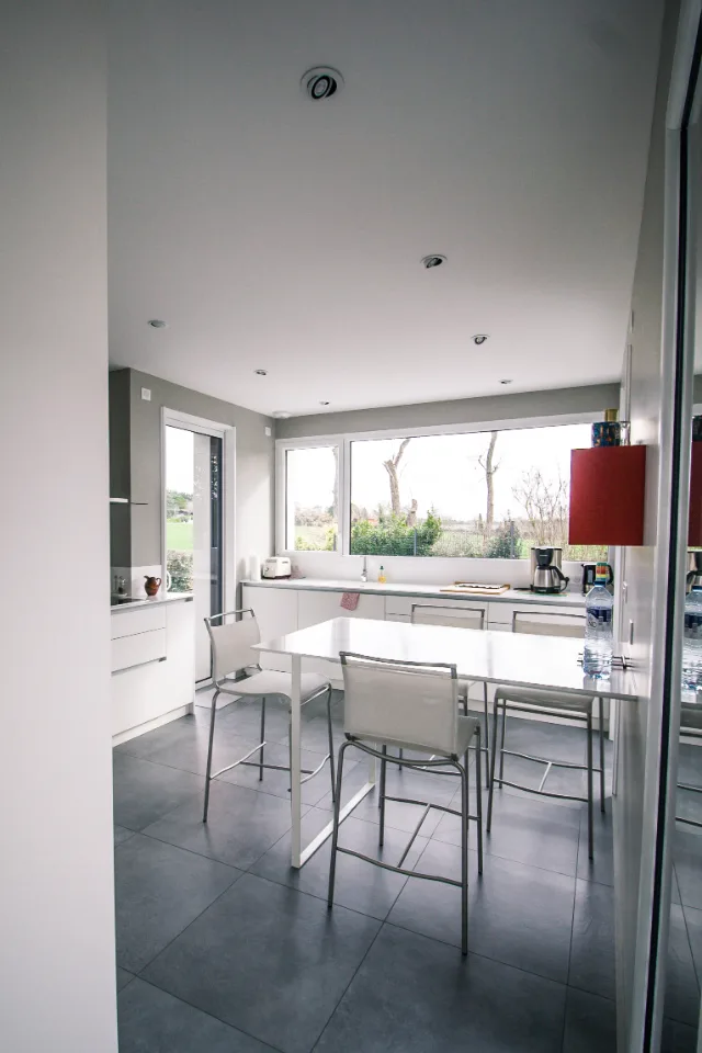 Cuisine blanche avec table haute, carrelage gris grand format, grande fenêtre et spots orientables