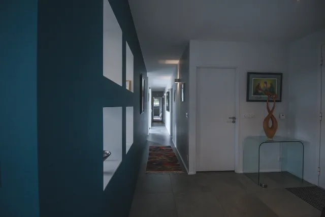 Couloir en pénombre avec mur bleu canard à niches, console en verre et appliques allumées