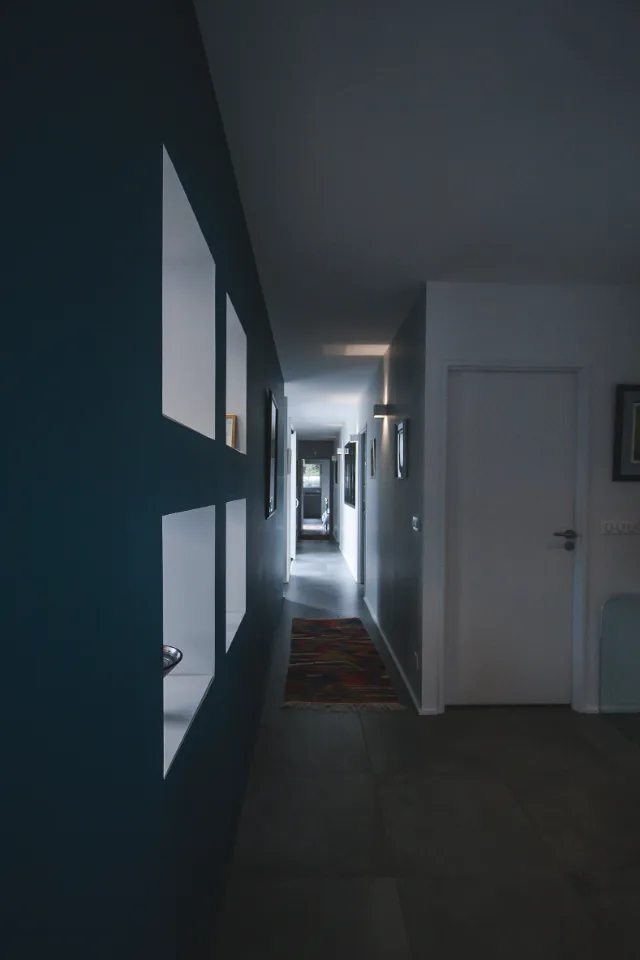 Long couloir sombre, niches blanches dans un mur bleu canard, appliques allumées et porte blanche