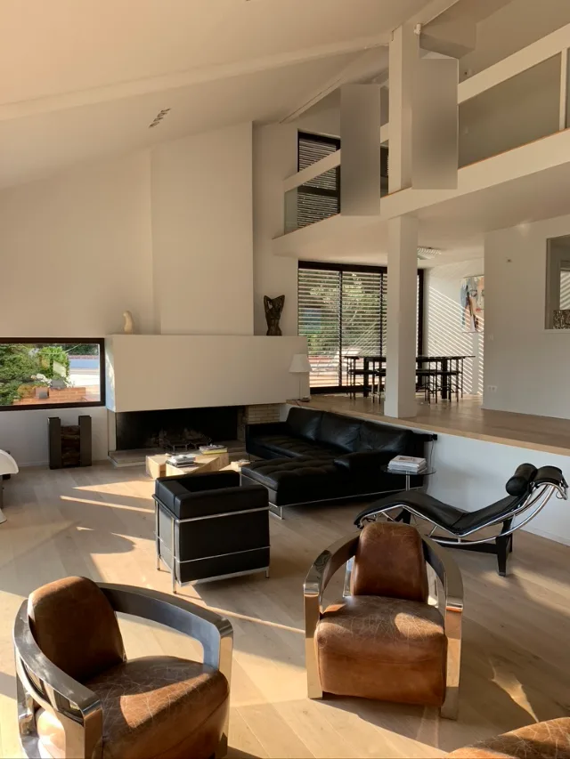 Séjour double hauteur avec cheminée blanche, fauteuils en cuir et mezzanine à garde-corps vitré
