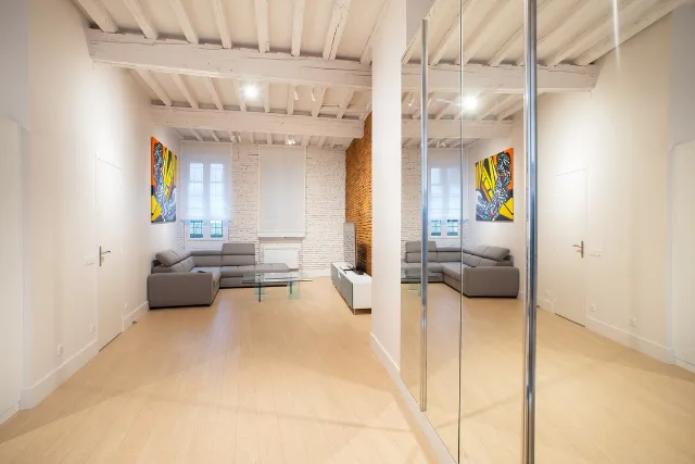 Séjour sous poutres peintes avec canapé d’angle gris, spots allumés et placard à portes miroir