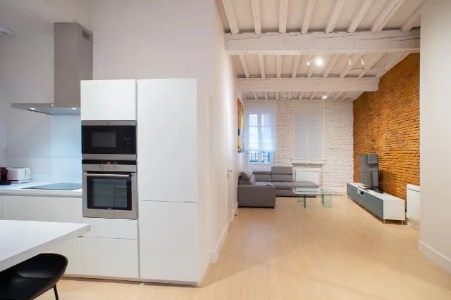 Cuisine blanche ouverte sur un séjour sous poutres, colonne four et micro-ondes, hotte inox
