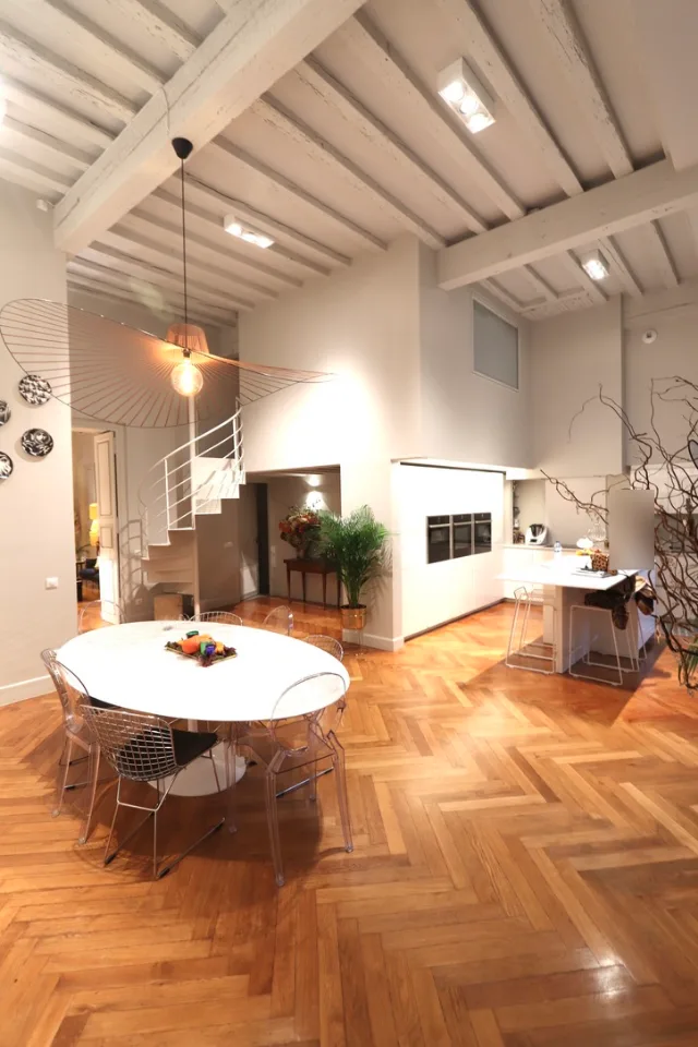 Grande pièce sous poutres, table ovale, suspension filaire, projecteurs en saillie et escalier hélicoïdal