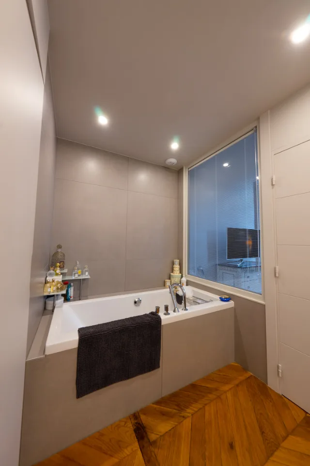 Salle de bains, baignoire dans un coffrage gris, spots encastrés et cloison vitrée à store
