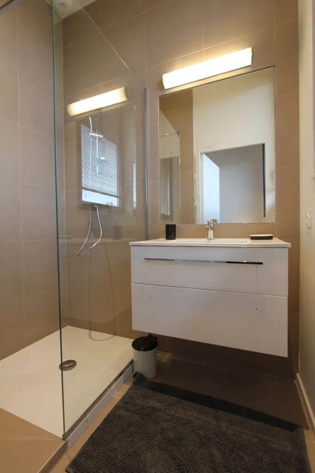 Salle d’eau avec douche à paroi vitrée, meuble vasque suspendu et applique allumée au-dessus du miroir