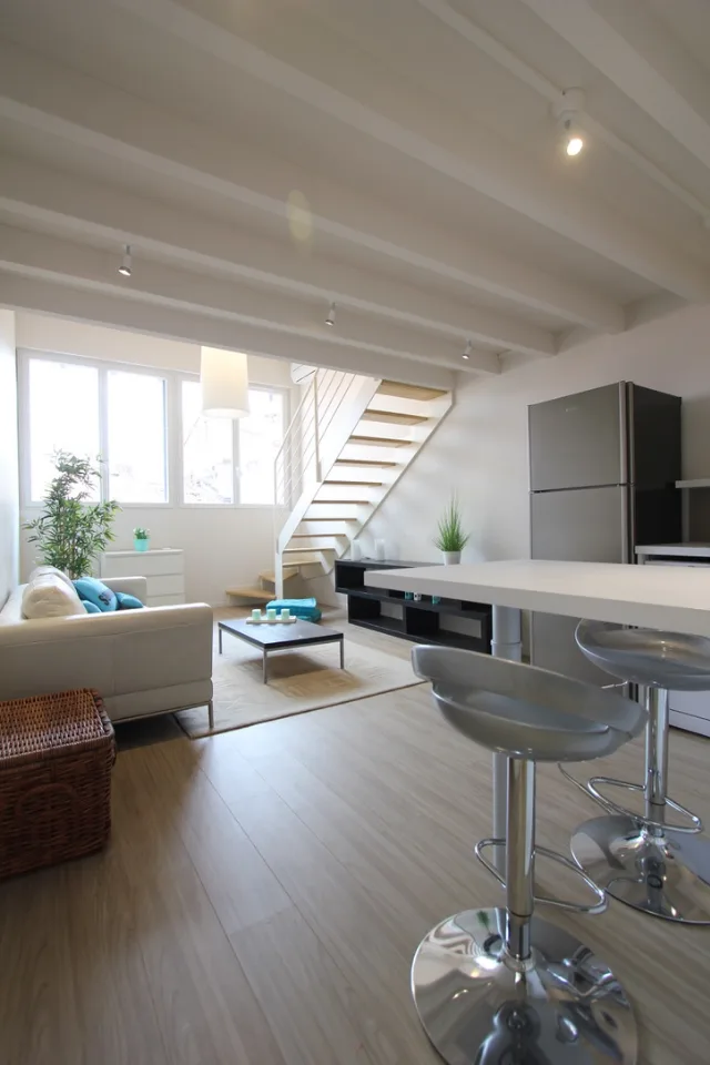 Séjour sous solives blanches avec spots orientables, escalier à marches en bois et bar à tabourets