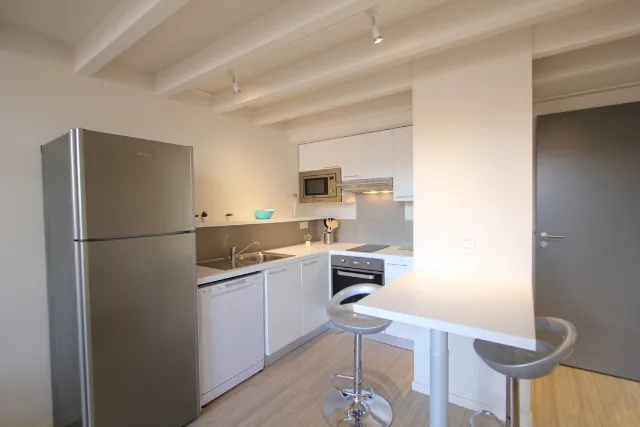 Cuisine blanche avec réfrigérateur inox, four, micro-ondes et hotte encastrés, bar à deux tabourets