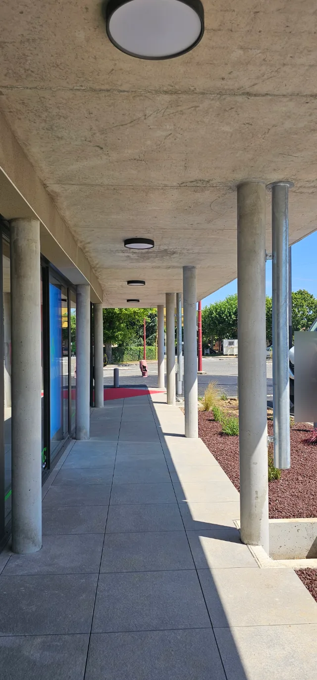 Galerie couverte en béton bordée de poteaux, plafonniers ronds en enfilade et massif de gravier rouge