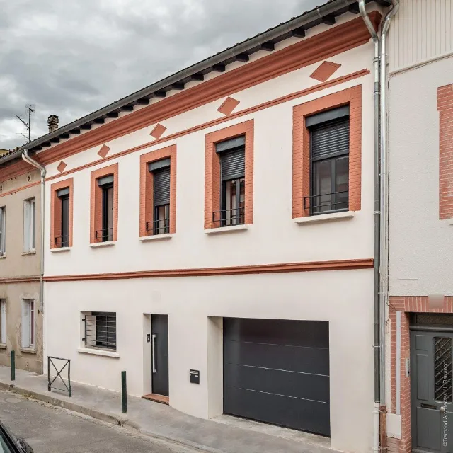 Façade sur rue enduite de blanc, encadrements de brique, volets roulants et porte de garage