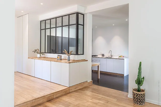 Pièce en parquet à deux niveaux, meuble bas sous verrière noire, cuisine blanche et spots encastrés