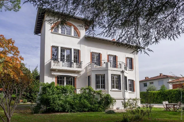 Maison blanche à tour carrée, encadrements de brique, balcons en fer forgé et jardin arboré