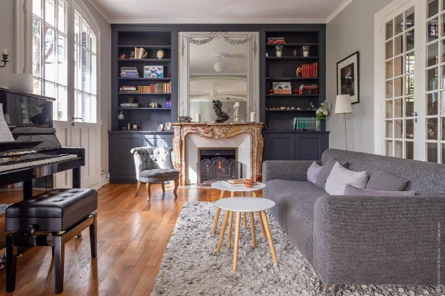 Salon avec cheminée en marbre allumée, bibliothèques gris-bleu, piano noir et canapé gris