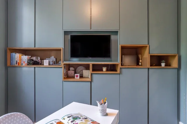 Pièce avec mur de rangements gris-bleu, niches en chêne, téléviseur encastré et bureau blanc