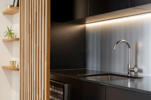Kitchenette noire, crédence inox éclairée par un ruban LED sous les meubles hauts
